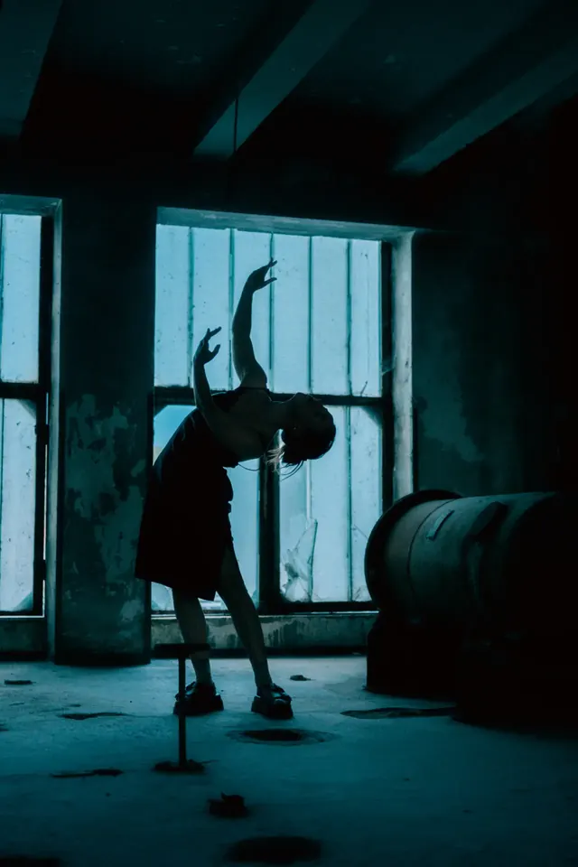 Dancer arching backwards in front of a factory window, teal tones